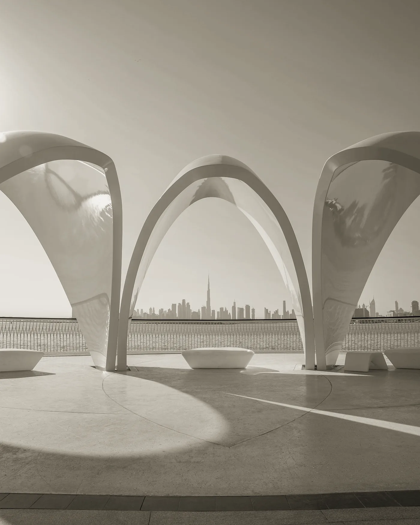 Arcos brancos de um pavilhão à beira-mar em Dubai, com o horizonte da cidade ao fundo, sob luz forte do sol.